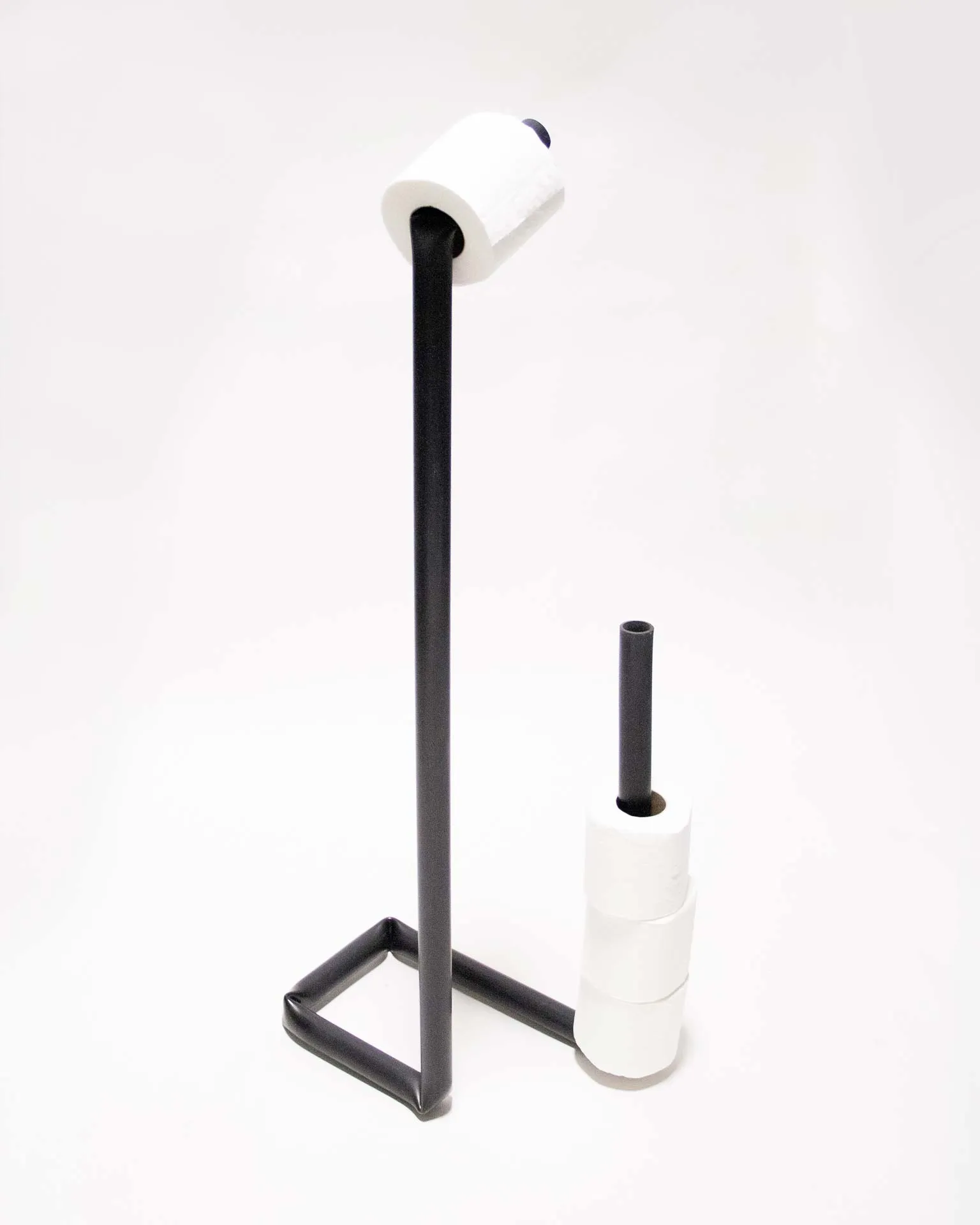 A toilet paper holder created by Jannis Schäfer Studio, made from a reclaimed PVC tube. The design features a black, L-shaped frame with one vertical rod holding a roll of toilet paper at the top and a second vertical rod holding multiple spare rolls. This object is part of a series of upcycled, recycled, and transformed designs.