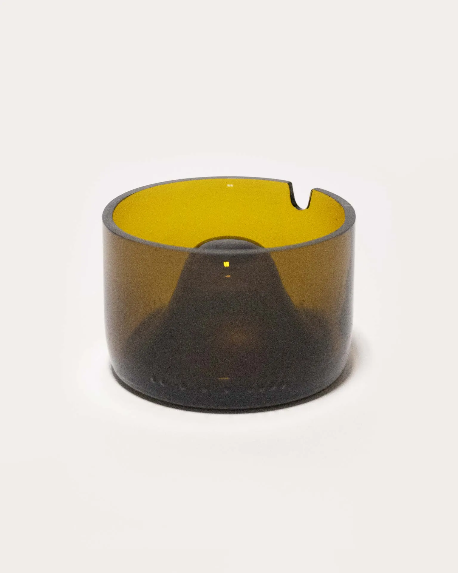 "An amber-colored cylindrical object made from upcycled glass, featuring a central dome and a small notch on the rim. The piece, designed by Jannis Schäfer Studio, is inspired by the Copenhagen ashtray by André Ricard and belongs to a series titled 'Derivatives'. The object highlights the use of reclaimed materials.”