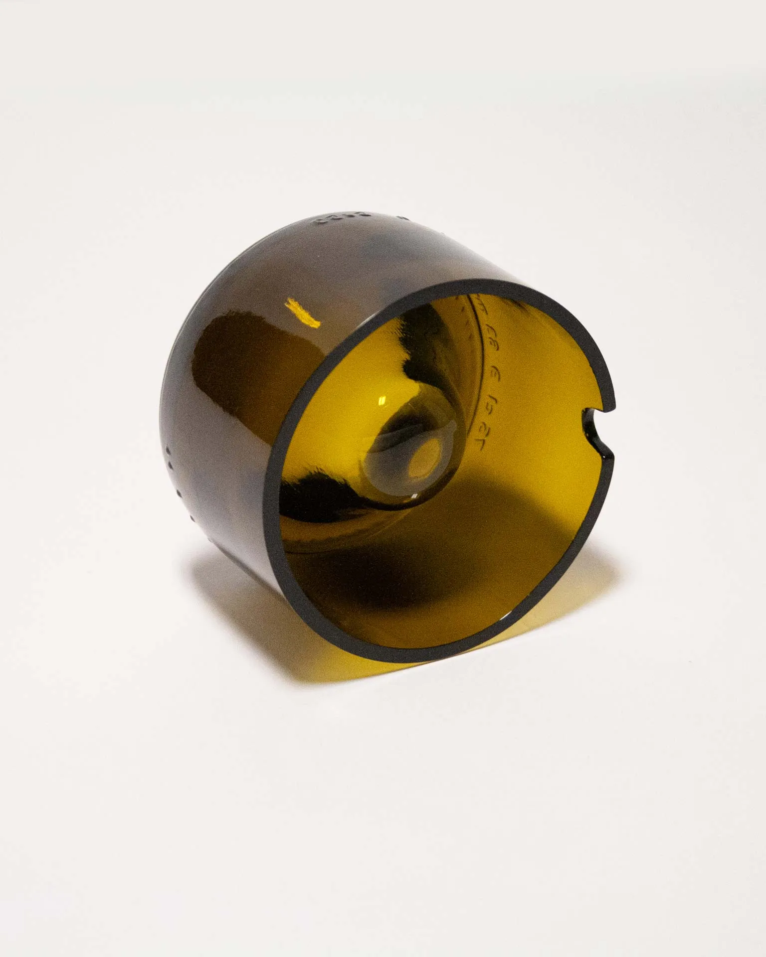 "An amber-colored cylindrical object made from upcycled glass, featuring a central dome and a small notch on the rim. The piece, designed by Jannis Schäfer Studio, is inspired by the Copenhagen ashtray by André Ricard and belongs to a series titled 'Derivatives'. The object highlights the use of reclaimed materials.”