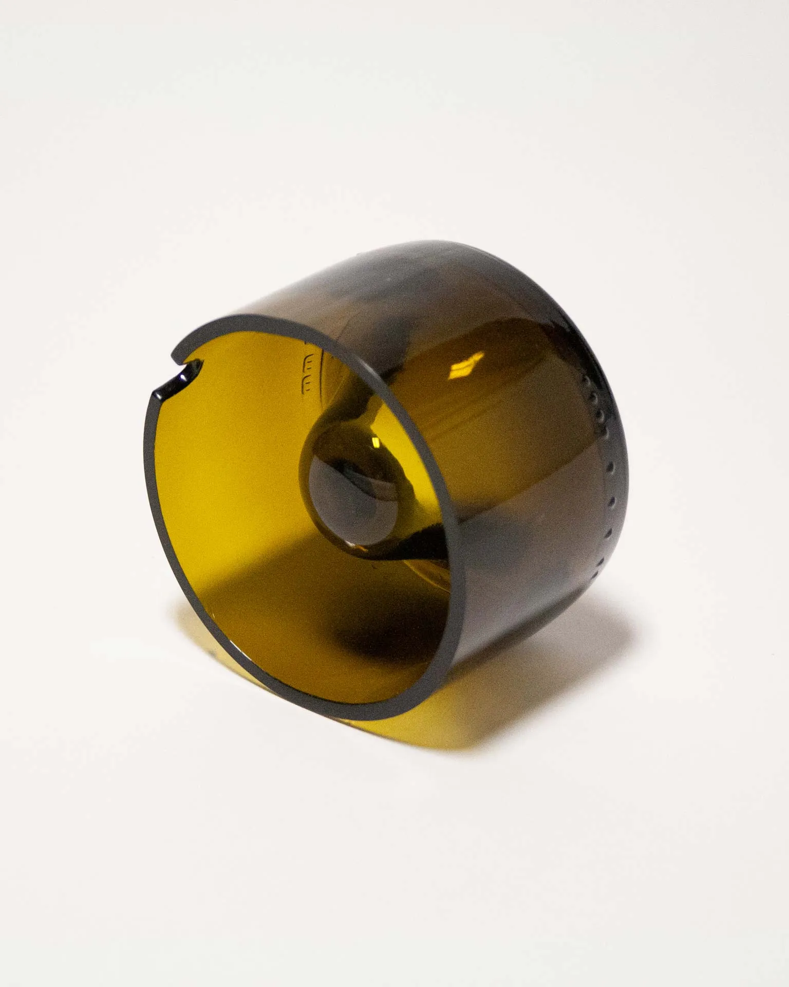 "An amber-colored cylindrical object made from upcycled glass, featuring a central dome and a small notch on the rim. The piece, designed by Jannis Schäfer Studio, is inspired by the Copenhagen ashtray by André Ricard and belongs to a series titled 'Derivatives'. The object highlights the use of reclaimed materials.”