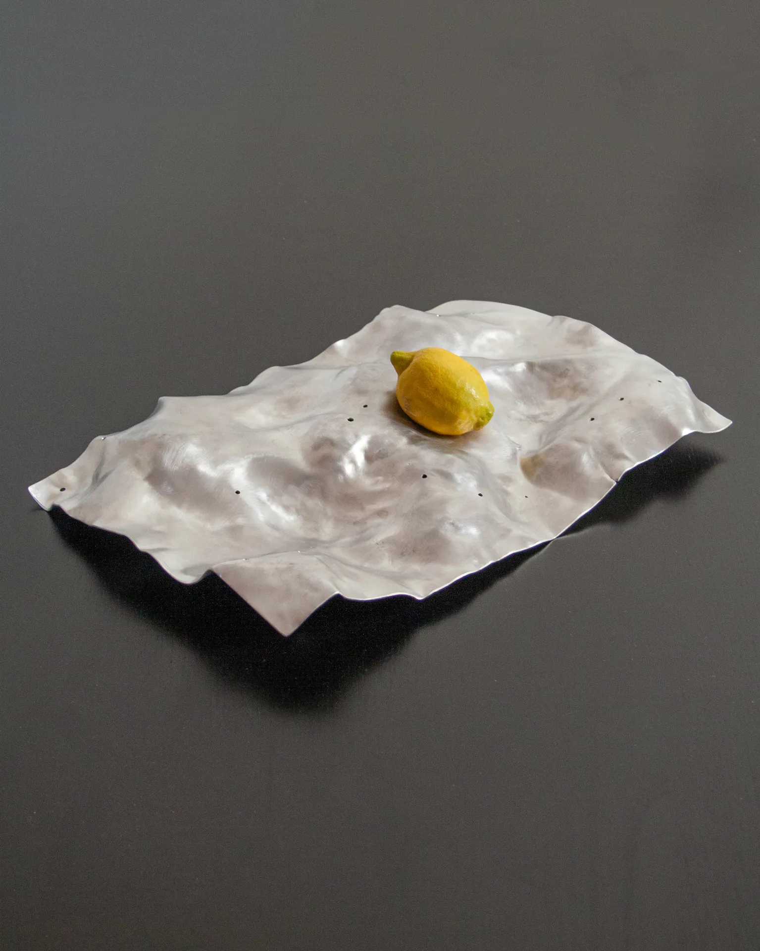 The 'Silver Platter,' created by Jannis Schäfer Studio, is made from a reclaimed aluminum motherboard tray. The design features a textured, undulating surface with a metallic finish. This upcycled, recycled, and transformed piece is part of a series of derivative designs.