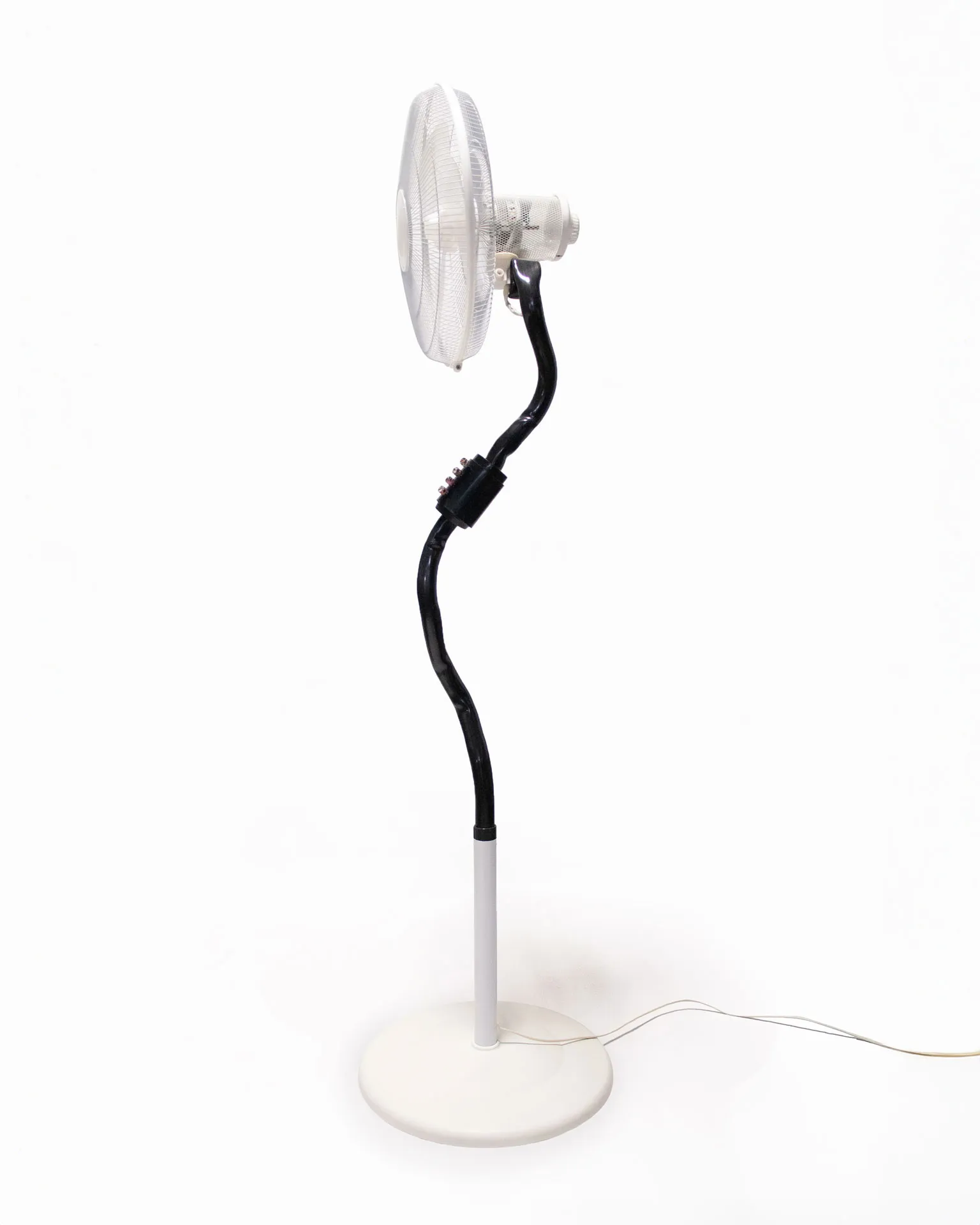 The 'Bent Pipe Blower,' created by Jannis Schäfer Studio, is a ventilator sculpture made from a reclaimed PVC tube and Precious Plastic samples as knobs and buttons. The design features a standing fan with a curved black pipe as the stand, a white circular base, and a clear fan head. This upcycled, recycled, and transformed piece is part of a series of derivative designs.