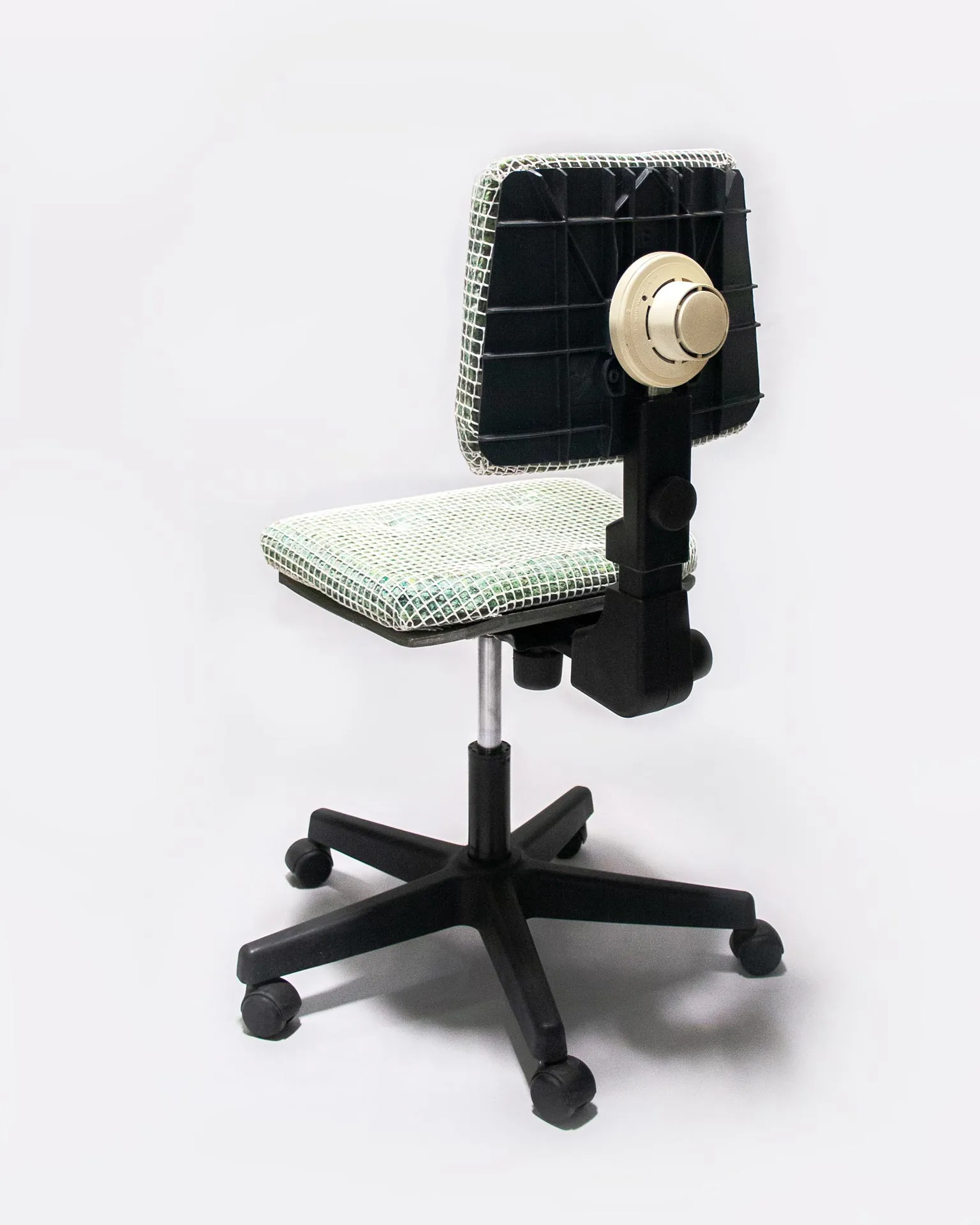 The 'Apollo Office Chair,' created by Studio Jannis Schäfer, is made from recycled insulation material, a plastic grid, and a smoke detector. The design features a cushioned seat and backrest covered in a green and white grid pattern, supported by a black five-wheel base with adjustable height. This upcycled, recycled, and transformed piece is part of a series of derivative designs.