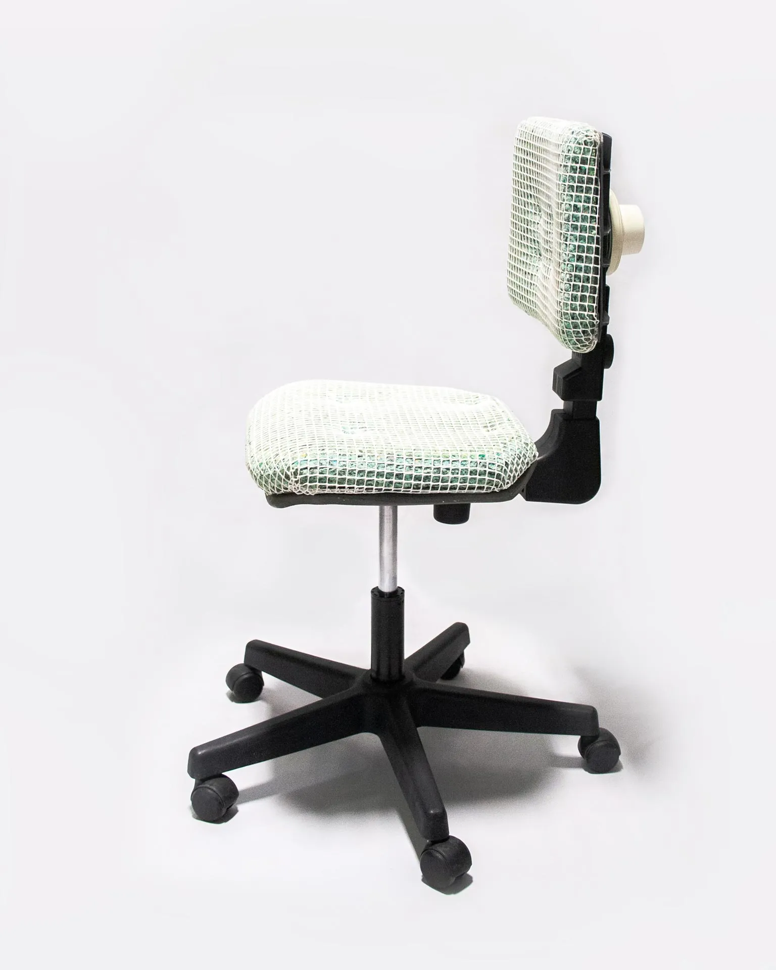 The 'Apollo Office Chair,' created by Studio Jannis Schäfer, is made from recycled insulation material, a plastic grid, and a smoke detector. The design features a cushioned seat and backrest covered in a green and white grid pattern, supported by a black five-wheel base with adjustable height. This upcycled, recycled, and transformed piece is part of a series of derivative designs.