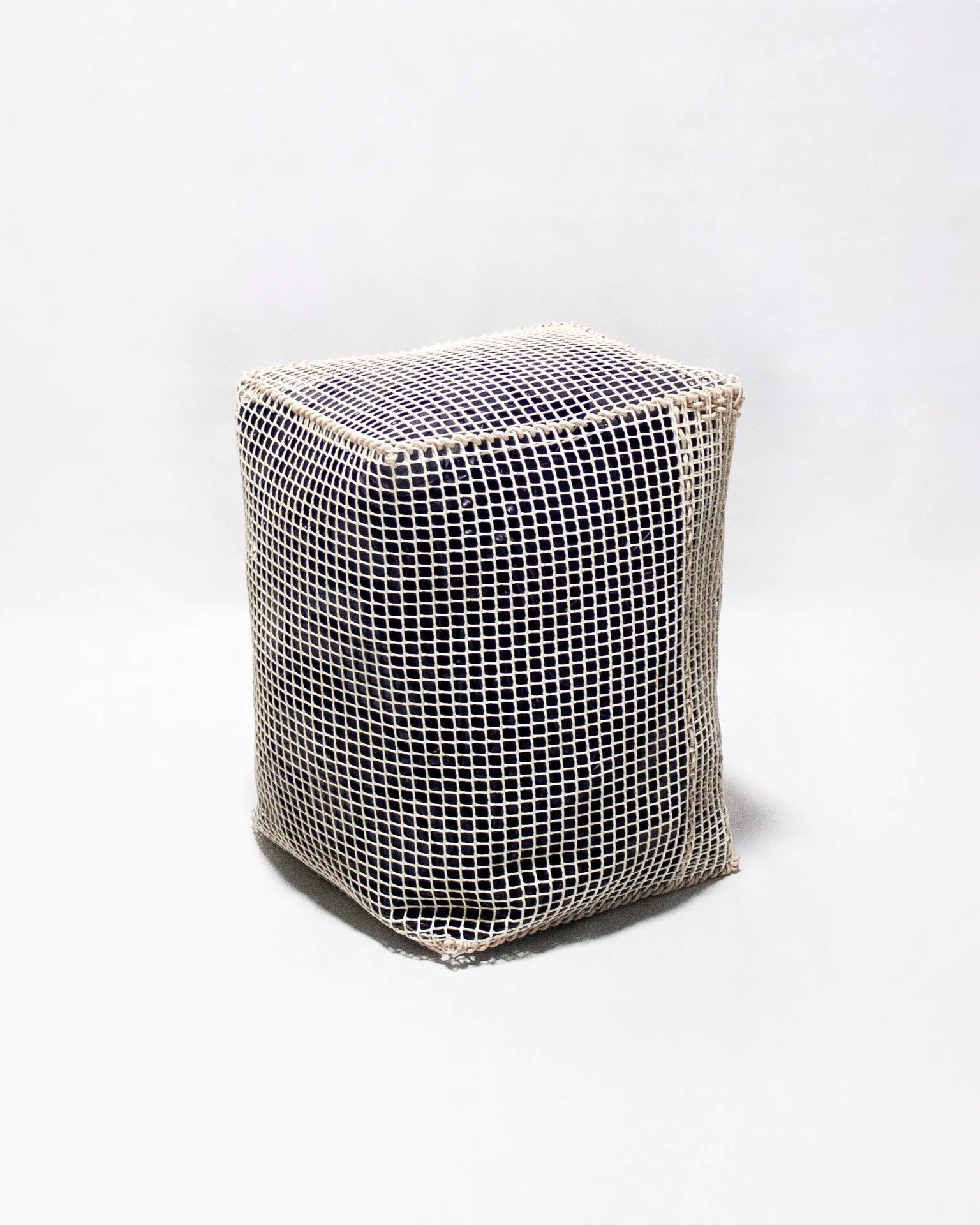 The 'Pinch Bag,' created by Studio Jannis Schäfer, is a pouf made from reclaimed anti-theft labels and a plastic grid. The design features a rectangular shape with a visible grid pattern overlaying the dark material inside. This upcycled, recycled, and transformed piece is part of a series of derivative designs.