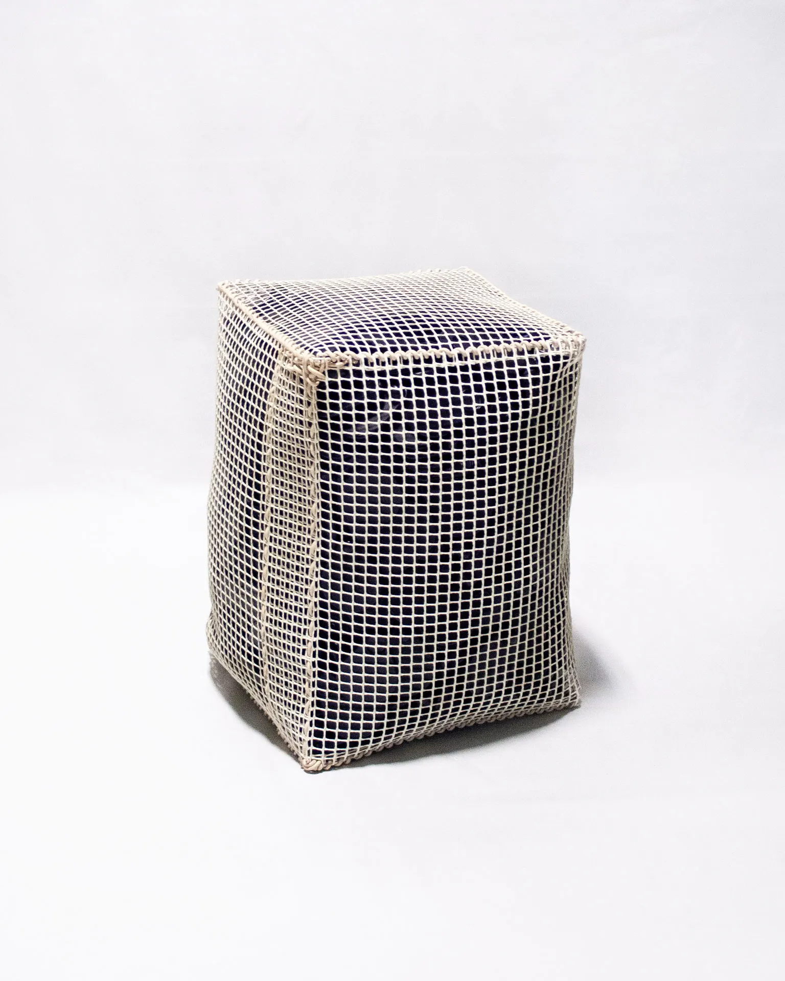 The 'Pinch Bag,' created by Studio Jannis Schäfer, is a pouf made from reclaimed anti-theft labels and a plastic grid. The design features a rectangular shape with a visible grid pattern overlaying the dark material inside. This upcycled, recycled, and transformed piece is part of a series of derivative designs.