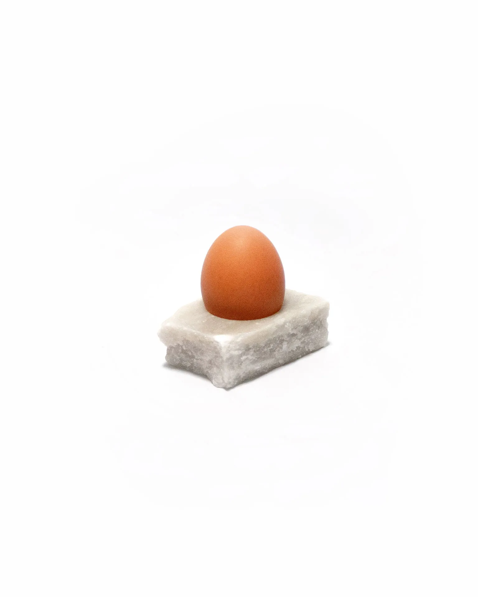 The 'Lithic Egg Cup,' created by Studio Jannis Schäfer, is made from a reclaimed marble tabletop. The design features a brown egg sitting on a small, rectangular marble base. This upcycled, recycled, and transformed piece is part of a series of derivative designs.