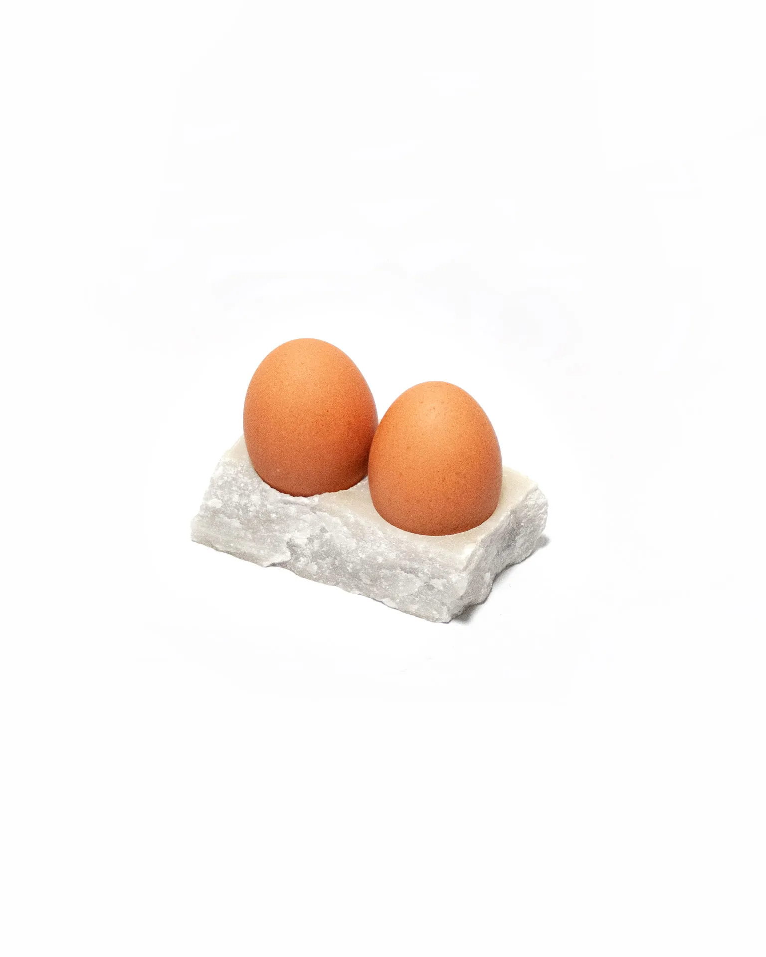 The 'Lithic Egg Cup,' created by Studio Jannis Schäfer, is made from a reclaimed marble tabletop. The design features a brown egg sitting on a small, rectangular marble base. This upcycled, recycled, and transformed piece is part of a series of derivative designs.