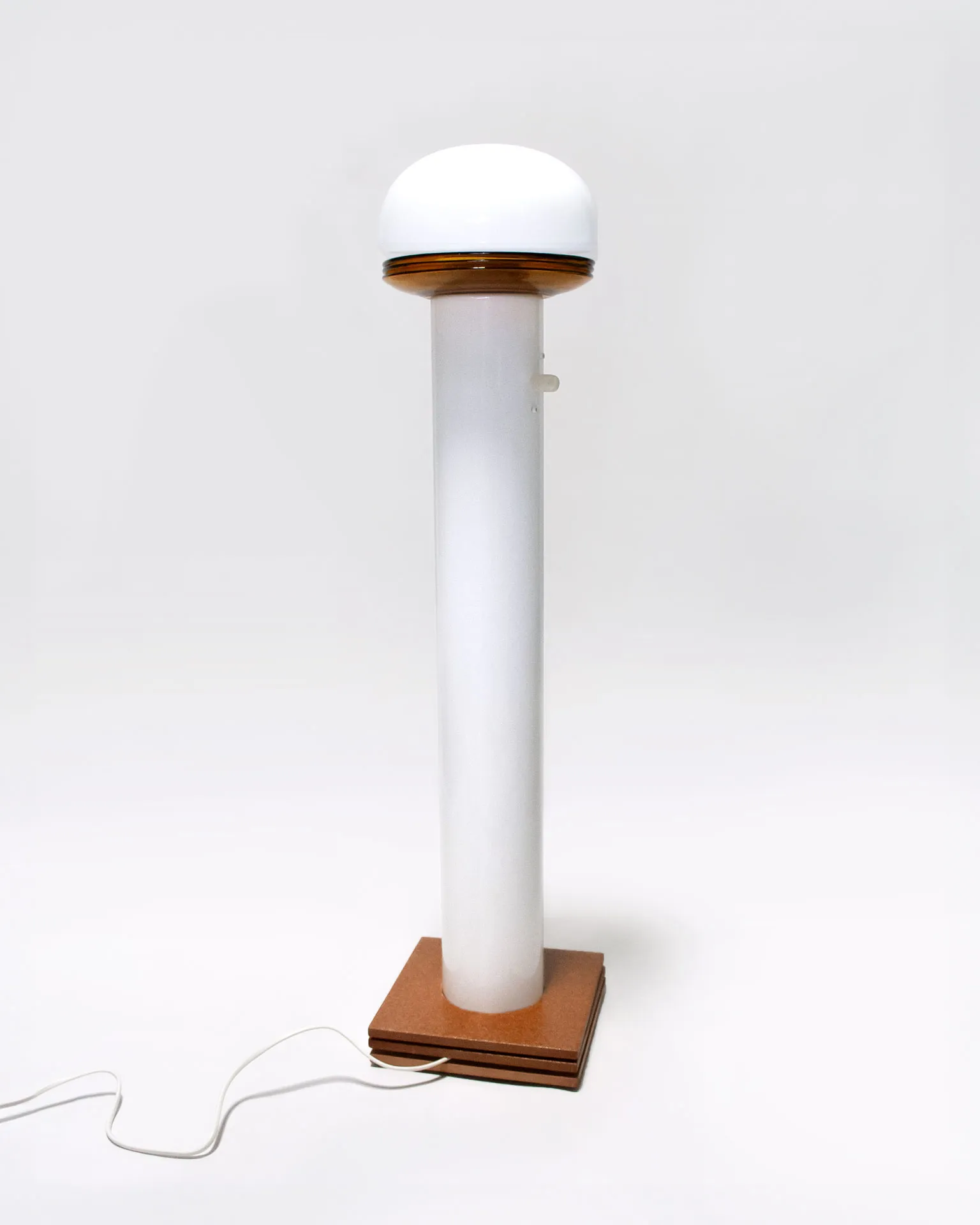 The 'Toadstool Lamp,' created by Jannis Schäfer Studio, is a dimmable lamp made from reclaimed tiles and a metal tube. The design features a white dome-shaped top resembling a toadstool cap, supported by a cylindrical base with a wooden square base. This upcycled, recycled, and transformed piece is part of a series of derivative designs.