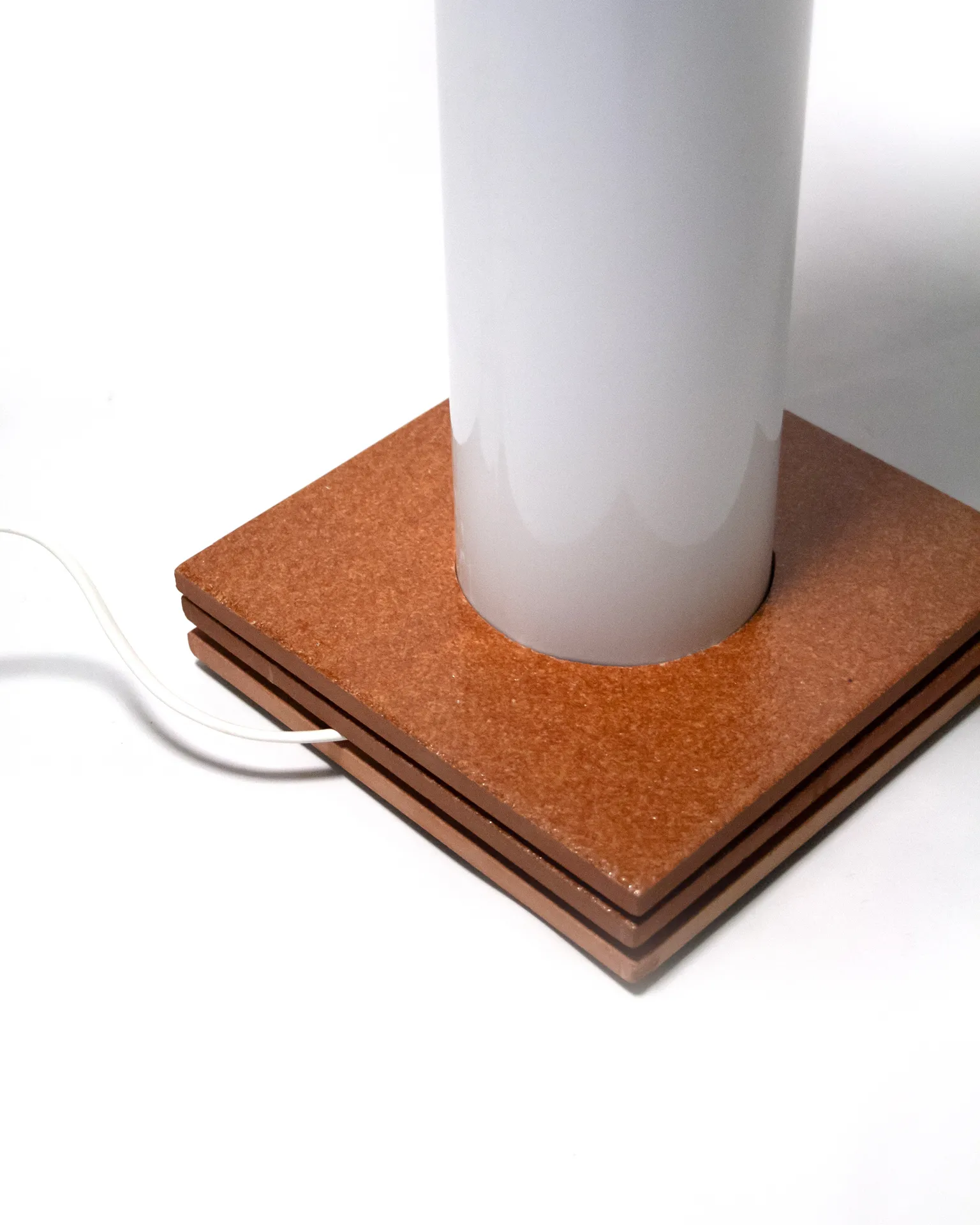 The 'Toadstool Lamp,' created by Jannis Schäfer Studio, is a dimmable lamp made from reclaimed tiles and a metal tube. The design features a white dome-shaped top resembling a toadstool cap, supported by a cylindrical base with a wooden square base. This upcycled, recycled, and transformed piece is part of a series of derivative designs.